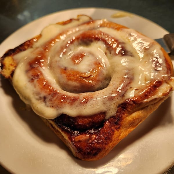 A large, warm Kountry Folks cinnamon roll with white icing on a white plate.