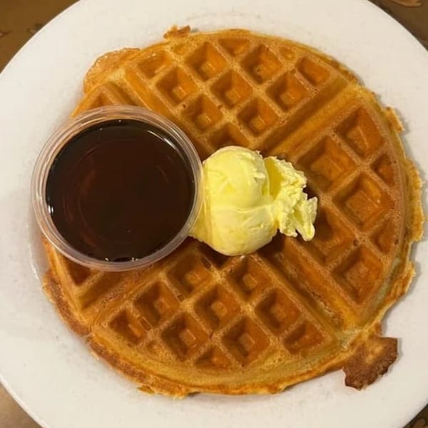 A golden waffle with a side of butter and a container of syrup on a white plate.
